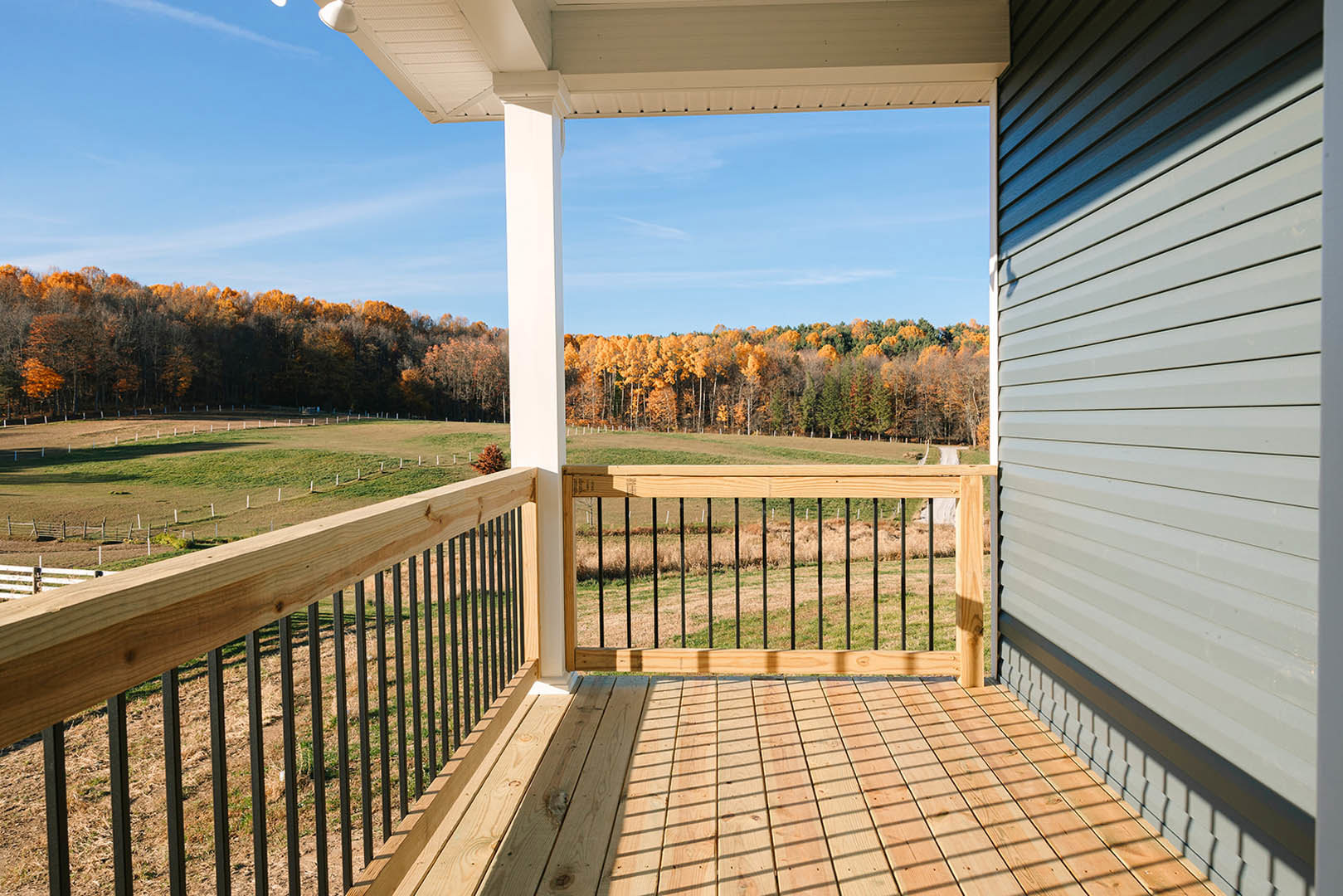 Wooden porch with black metal railing, white paneled ceiling, fenced deck overlooking grassy yard and trees in autumn.