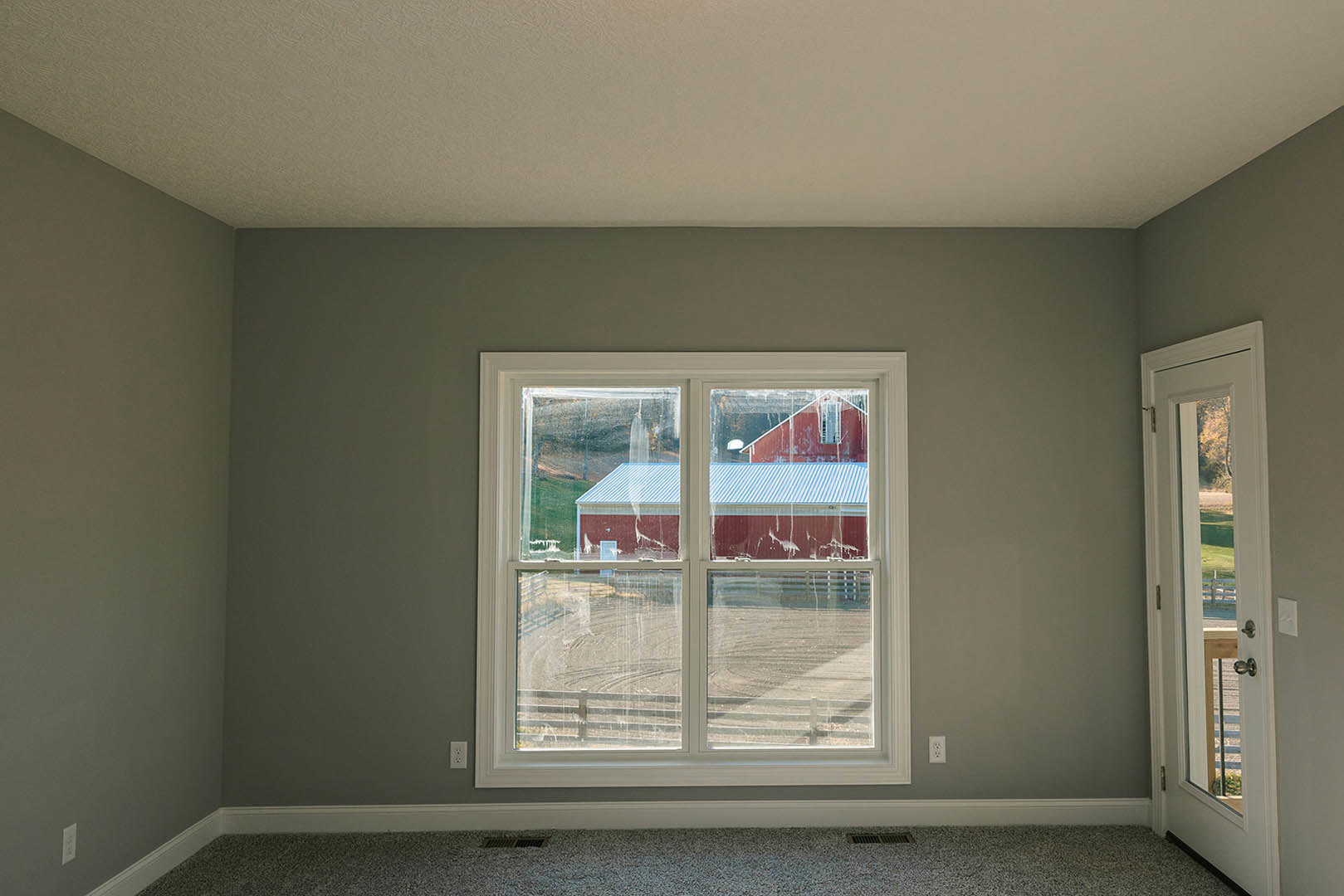 Large window with white trim in a plaster-walled room, open door leading to fenced yard, view of barn and red building outside, blue roof visible, beige carpet flooring