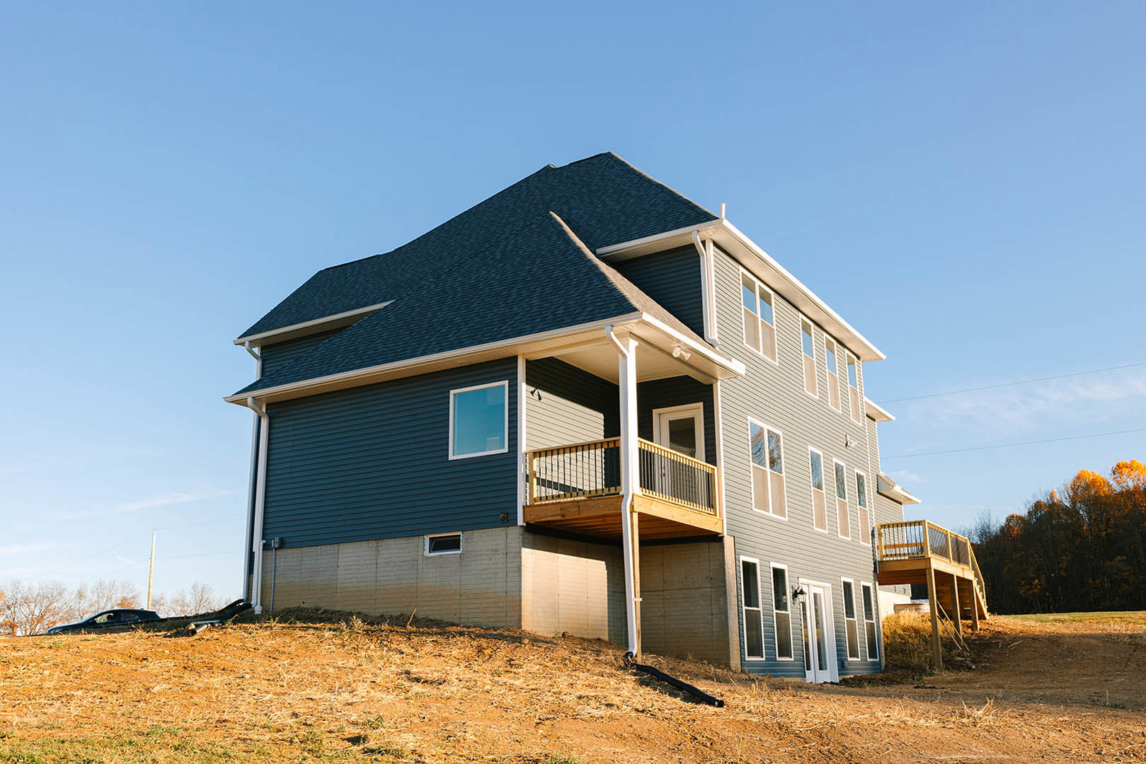 Two-story house with light siding, white-framed windows, and a wooden side deck with railing, surrounded by mature trees