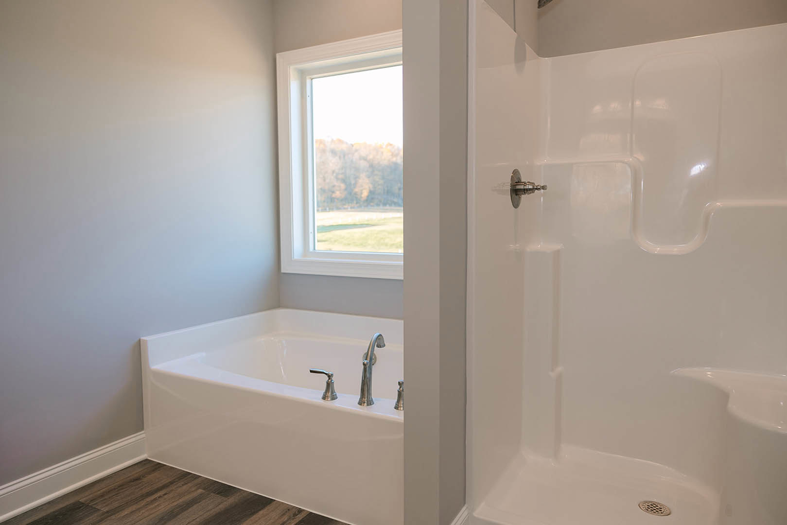 Bathroom with freestanding bathtub, silver faucet, wood flooring, glass-enclosed shower with chrome handle, large window overlooking trees, white tile walls