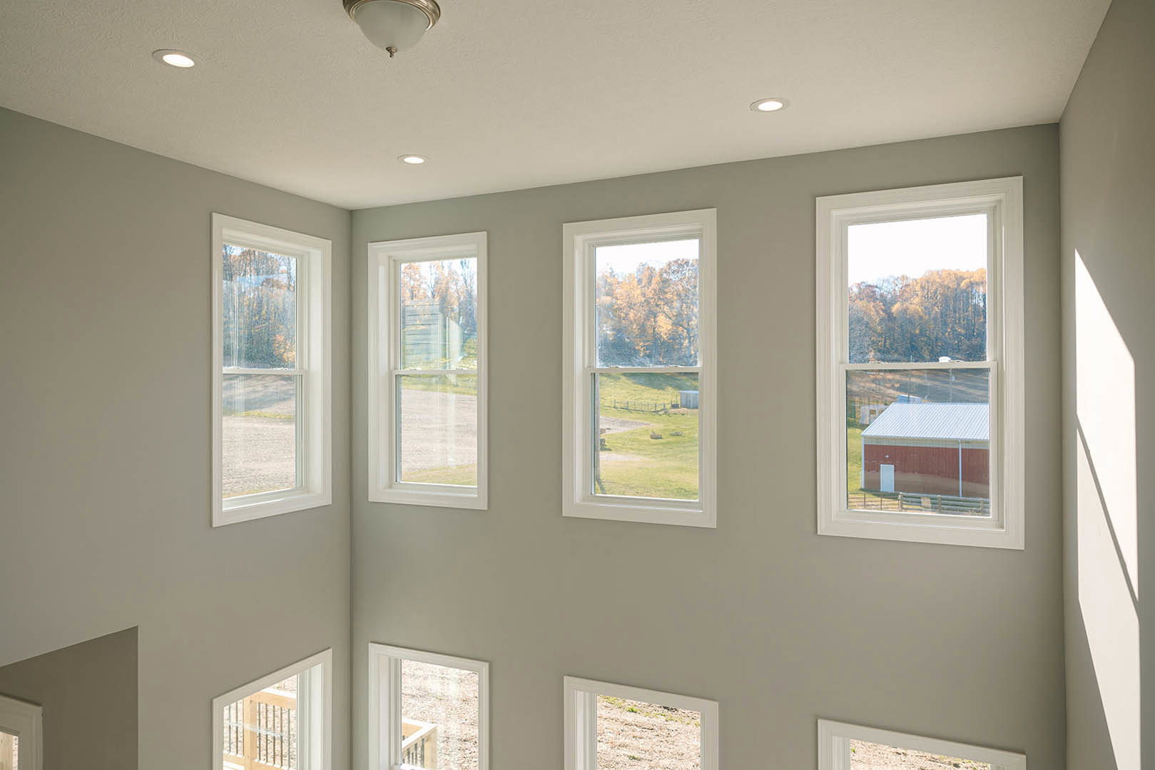 Spacious room featuring multiple large windows with views of fields, trees, and a distant farm; light-colored walls, ceiling, and a modern ceiling light fixture.