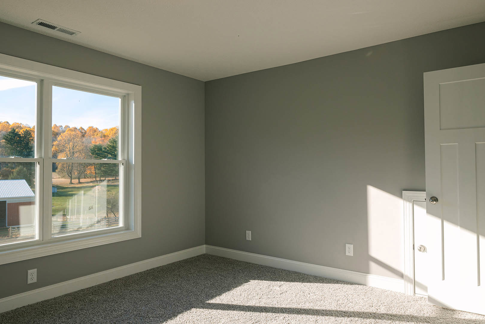 Bedroom with beige carpet, white walls, large window overlooking trees and neighboring house, white door, ceiling vent, and recessed lighting.