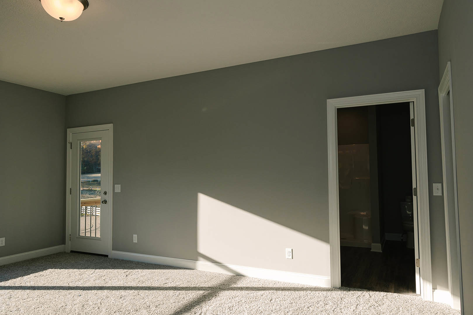 White-walled room with two doors, one opening to a bathroom and the other revealing a fenced yard with grass, white carpet flooring, recessed ceiling light fixture
