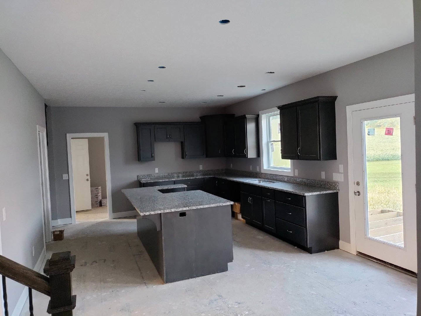 Black kitchen cabinets and countertops, white door with silver knob, wooden staircase railing, view through door to grassy field, light-colored flooring, stacked boxes near entry.