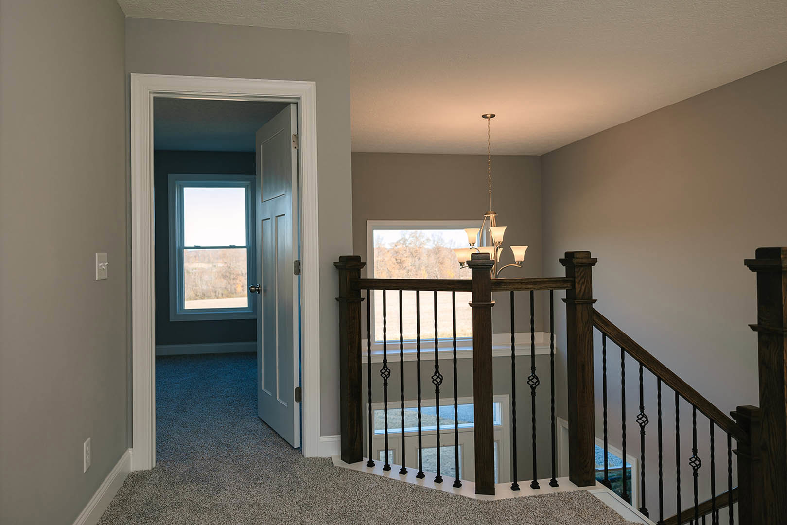 Wood staircase with metal railing leads to open doorway, light hardwood floors, white walls, large window overlooking grassy field, modern chandelier hanging above.
