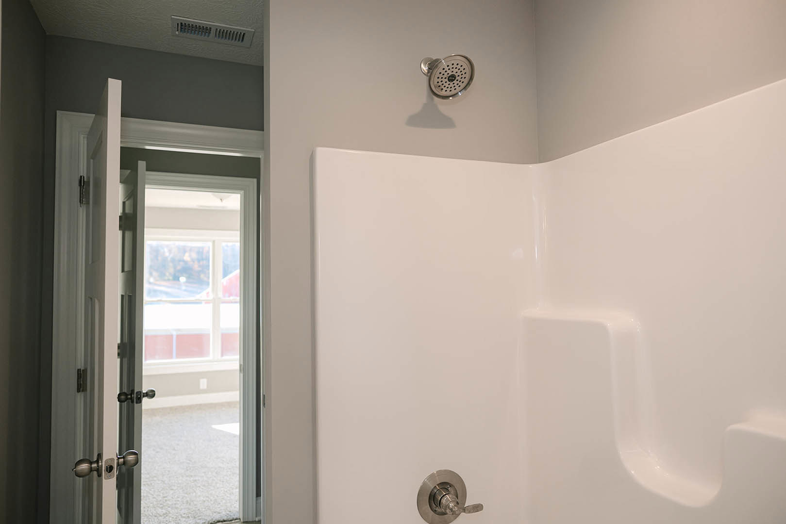 White bathtub with chrome faucet and shower head, adjacent to a wall vent and open door revealing a window, plaster walls and ceiling, modern bathroom finishes.