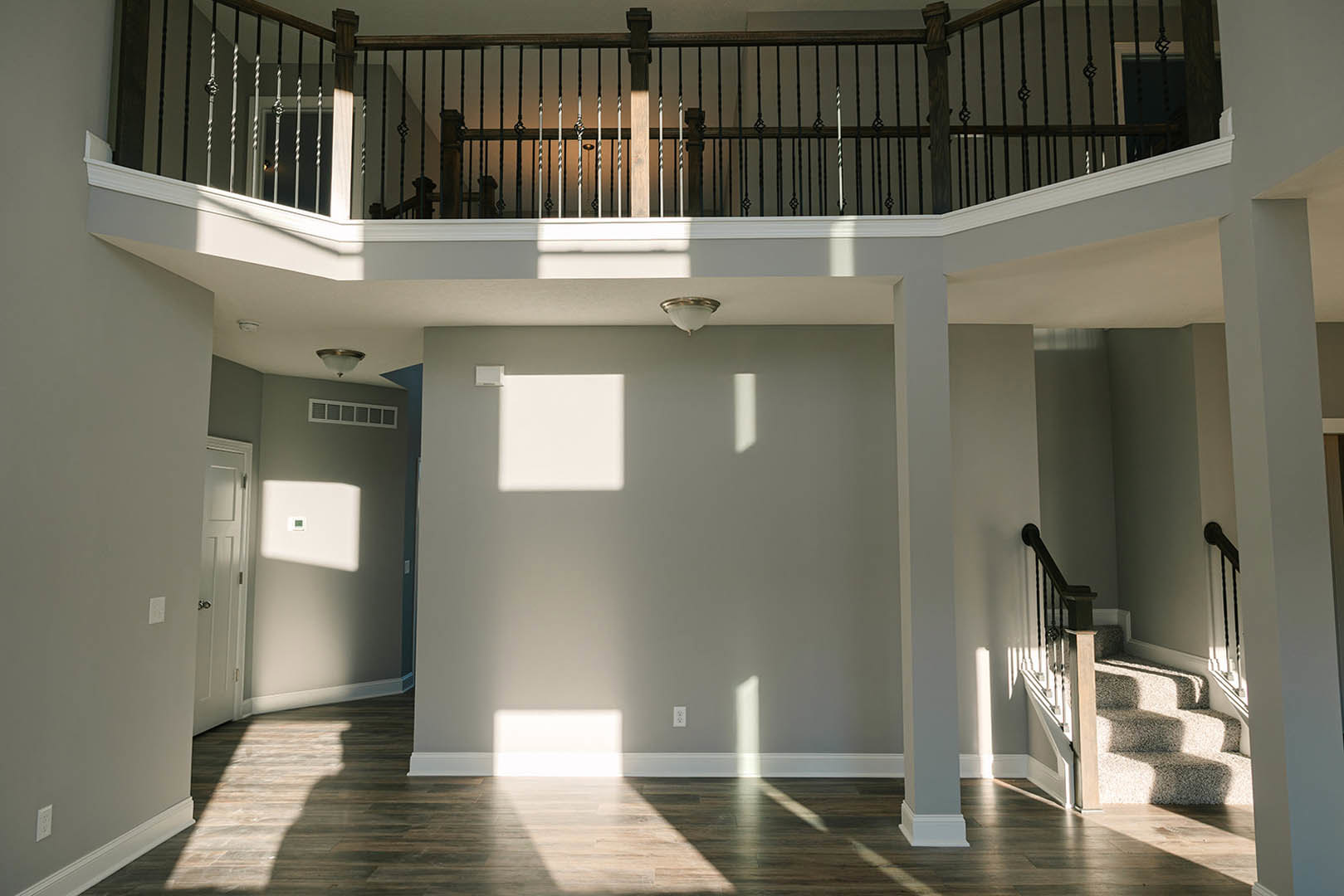 Open-concept room featuring a wood floor, white walls, modern staircase with metal handrail, close-up of a light fixture, and a white door with a silver handle.