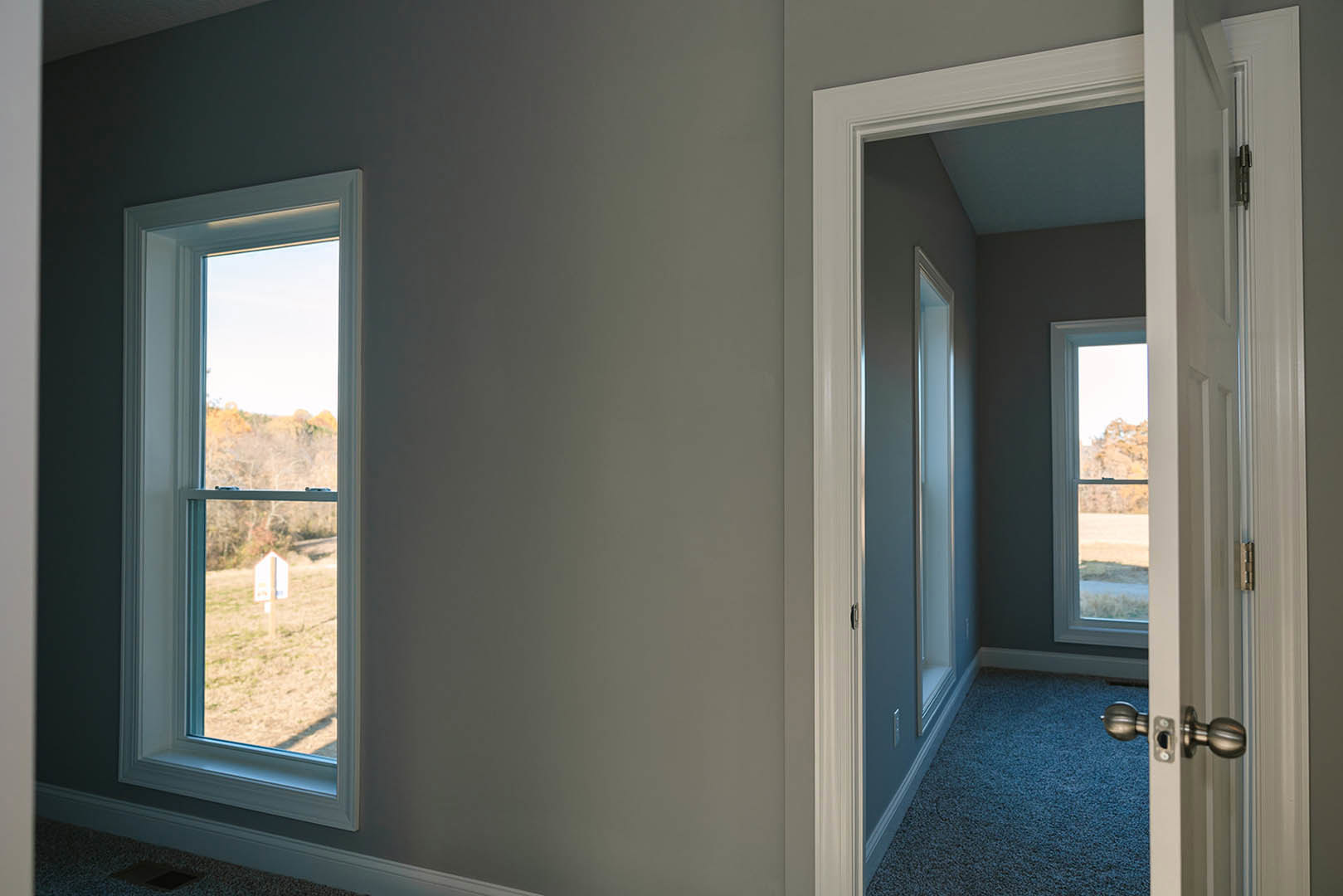 Hallway with light-colored walls, two large windows overlooking a grassy field, close-up of a modern door handle, window blinds partially drawn, daylight streaming in