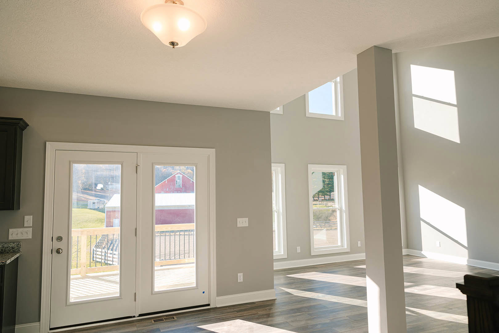 Double glass-paneled doors and windows in a room with light plaster walls, wood flooring, and a two-light ceiling fixture; window view includes a fence and trees.