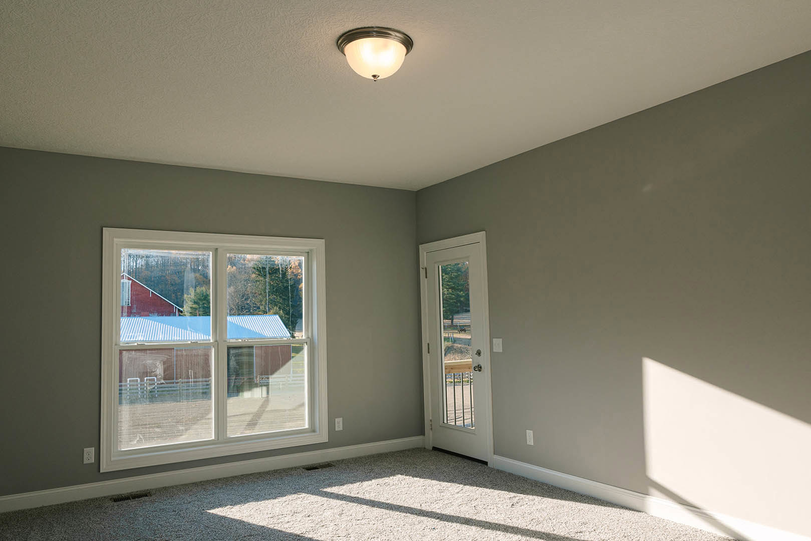 Spacious room featuring white plaster walls, large windows with views of a barn and blue roof, wooden door, ceiling-mounted light fixture, and neutral carpet flooring.