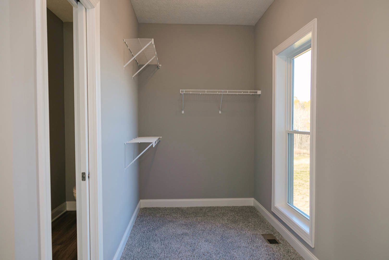 White built-in shelves on plaster wall, large window with outdoor view, wood floor transitioning to carpet, white metal ceiling bar, minimalist interior with neutral finishes.