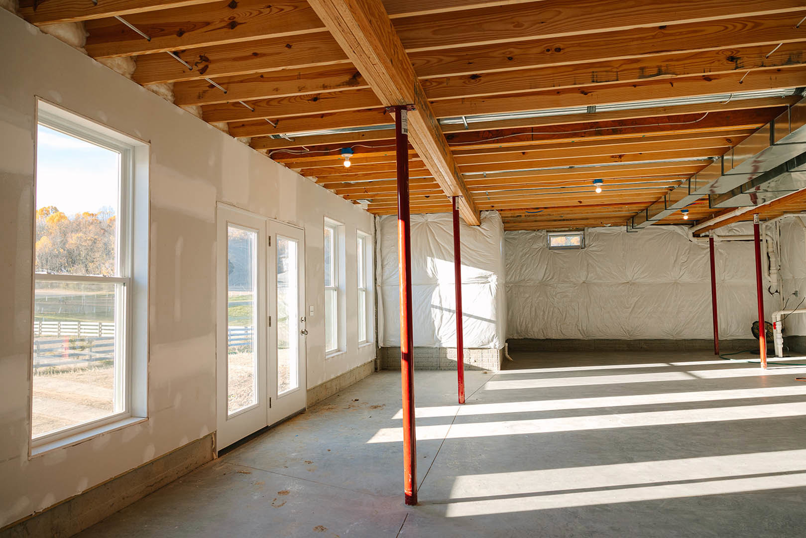 White-walled room with red structural poles, concrete floor, wooden ceiling beam, large window showing trees, and a dog standing indoors