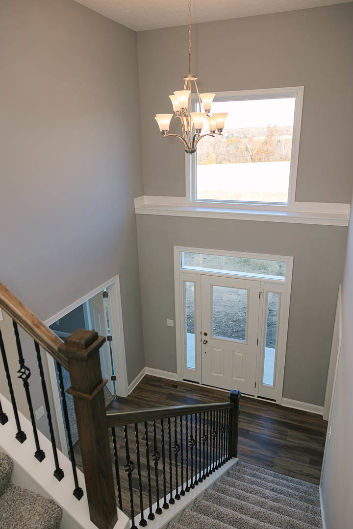 Wooden staircase with dark handrail and white balusters, white door with glass panes, chandelier hanging in front of large window, crown molding and plaster walls in entryway.