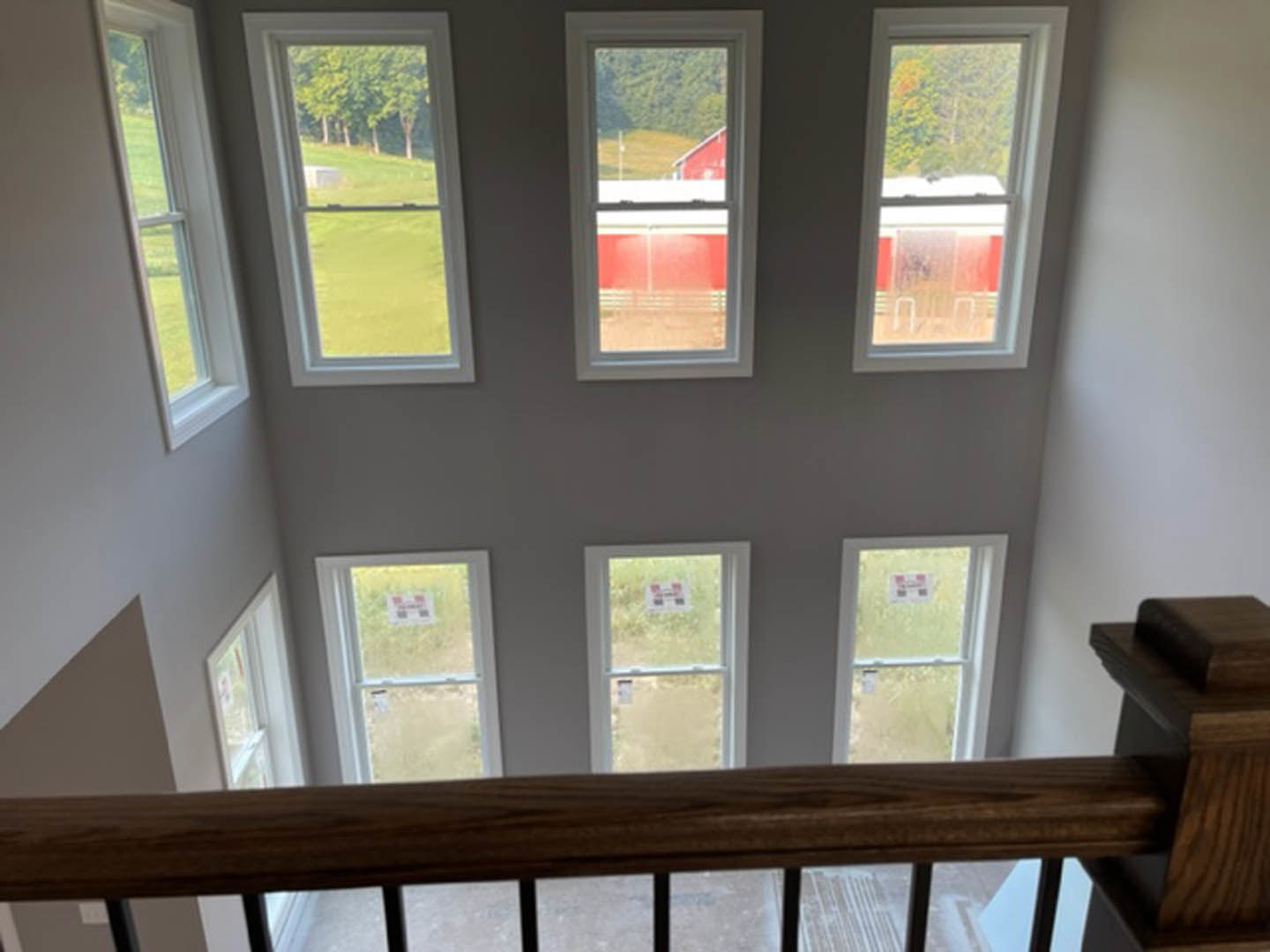 Wood staircase with horizontal railing, multiple large windows providing daylight, view of red barn and trees outside, window with sign, hardwood floors, white walls