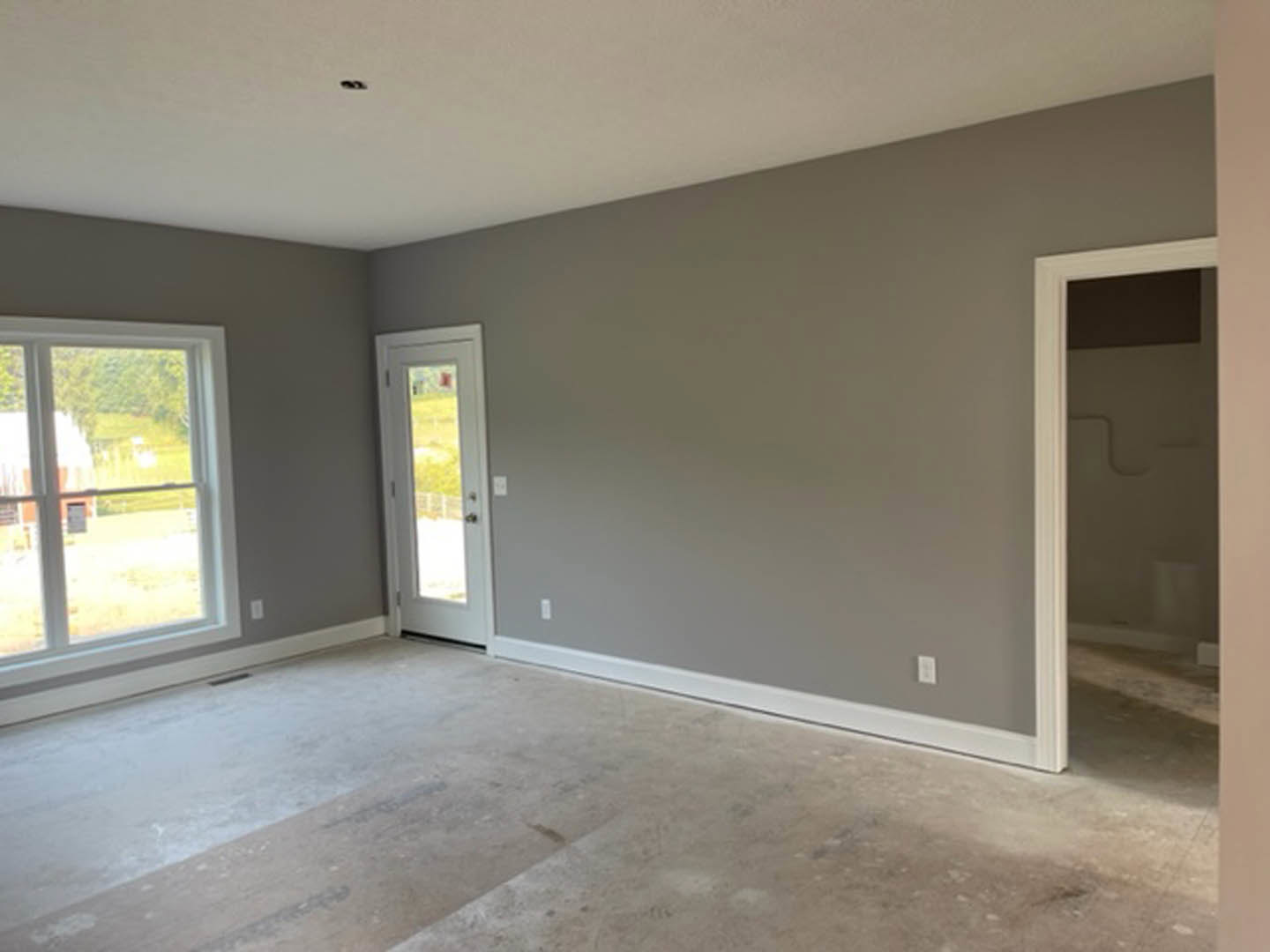 Bright room with concrete flooring, white trim, a white door with glass panels, and large windows framed in white overlooking a grassy field