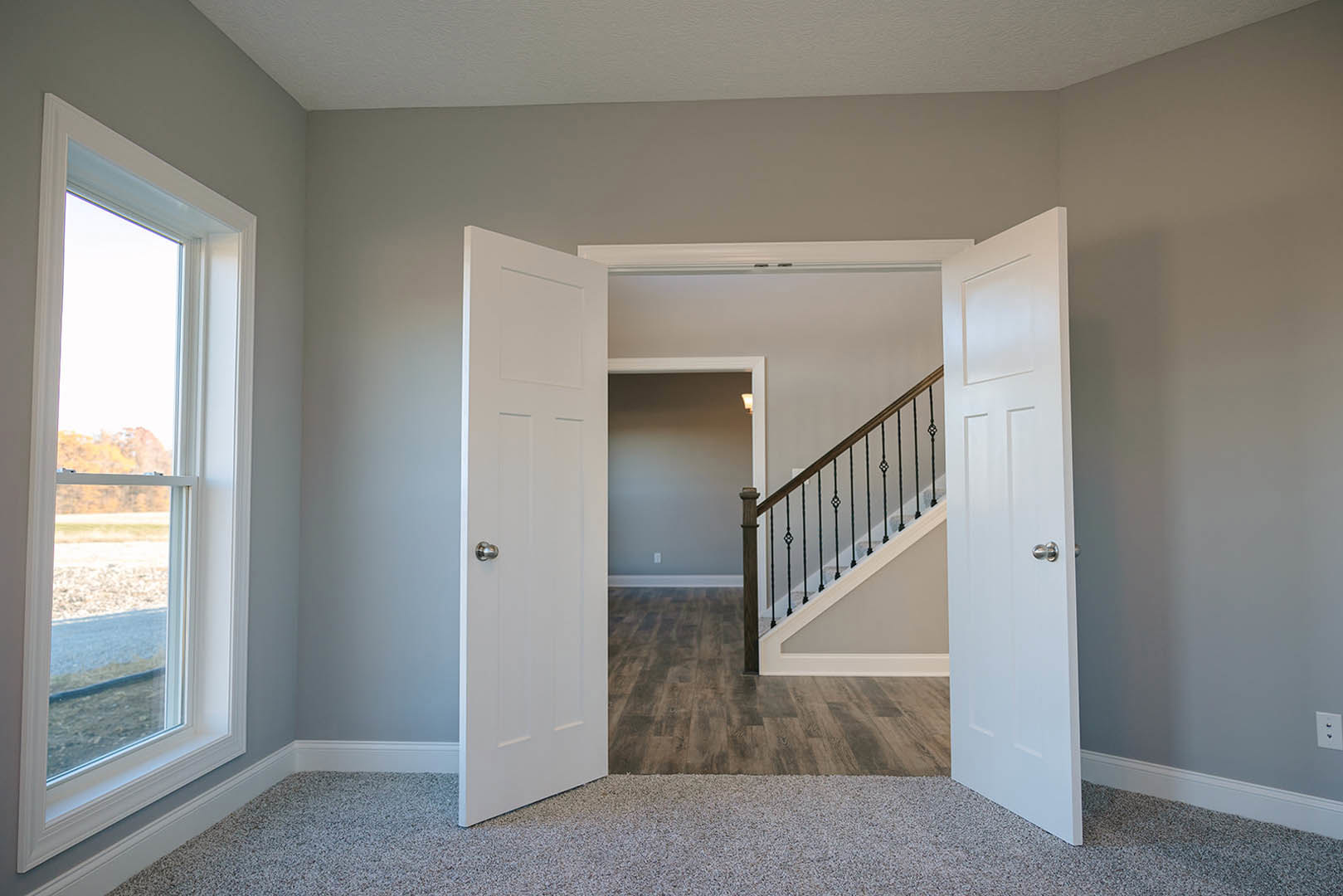 White-painted staircase with wooden treads, two paneled doors, beige carpet flooring, large window overlooking grassy field, smooth plaster walls with crown molding
