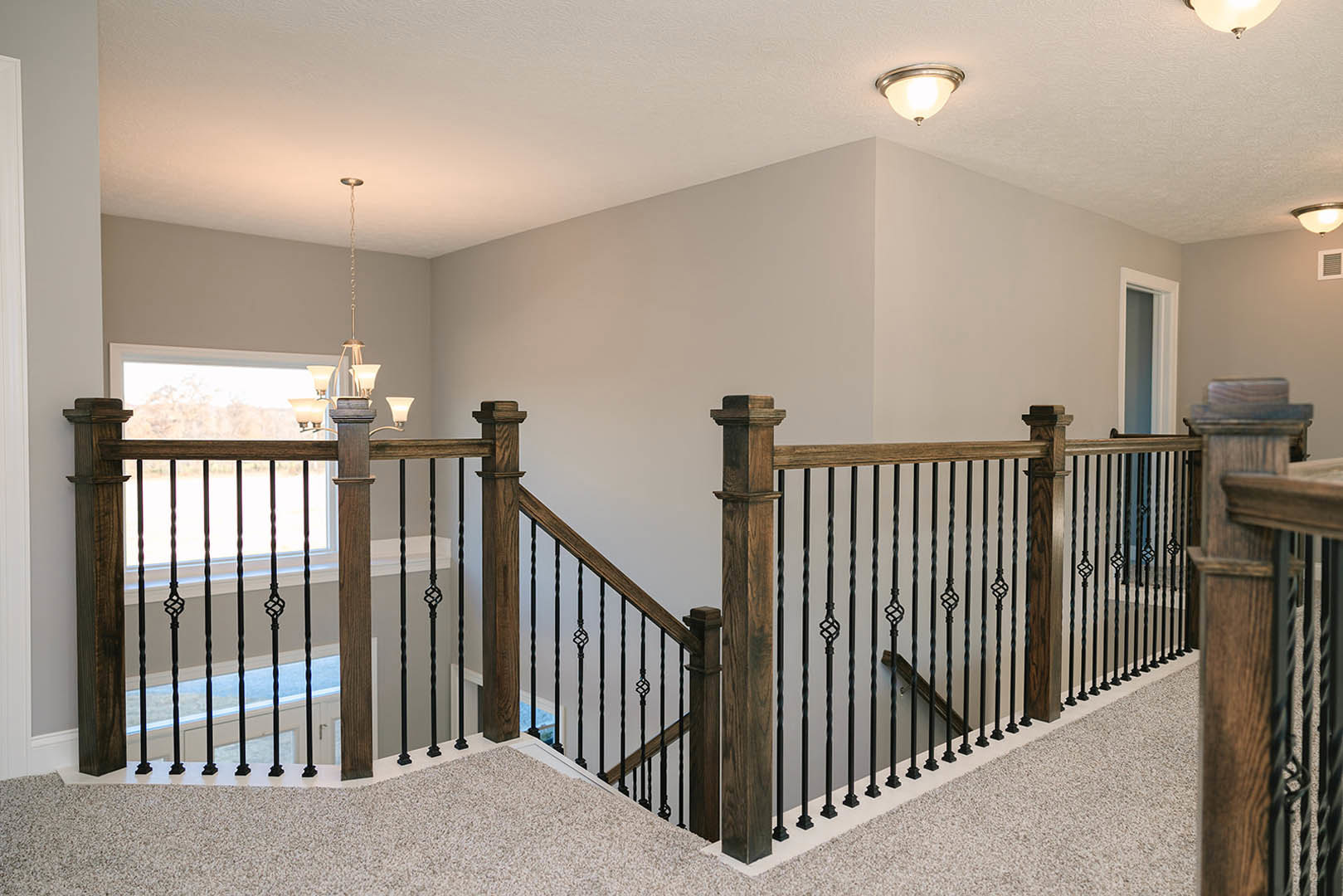 Wood staircase with black iron railing, illuminated by a ceiling light fixture, adjacent to white plaster walls and a closed door, with hardwood flooring throughout.