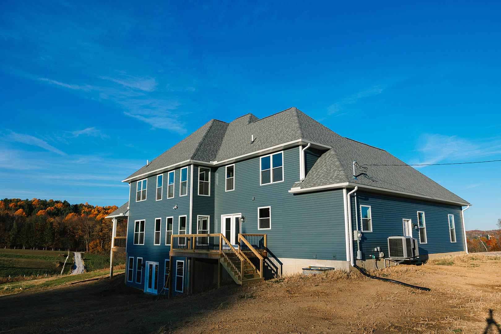 Two-story house with wooden deck and stairs, white siding, large windows, surrounded by trees with orange leaves under blue sky