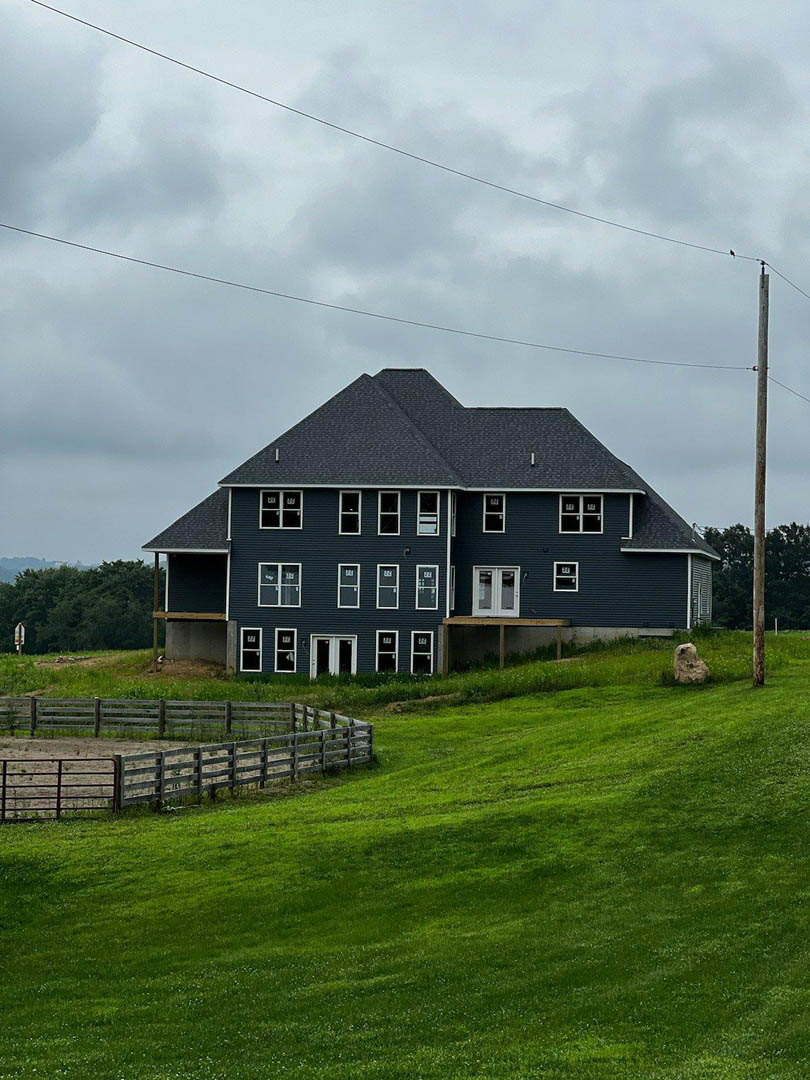 Two-story house with gray siding and gabled roof, surrounded by a wooden fence and grassy yard, mature trees in background, partly cloudy sky overhead