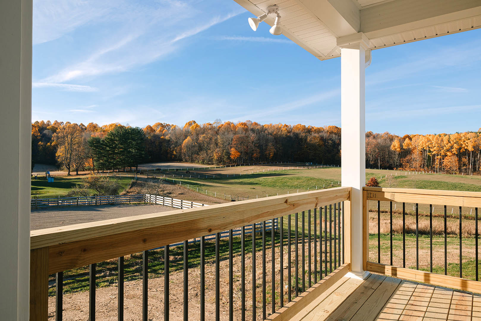 Wooden deck with black metal railings, white ceiling supported by columns, fenced yard bordered by tall trees with autumn foliage