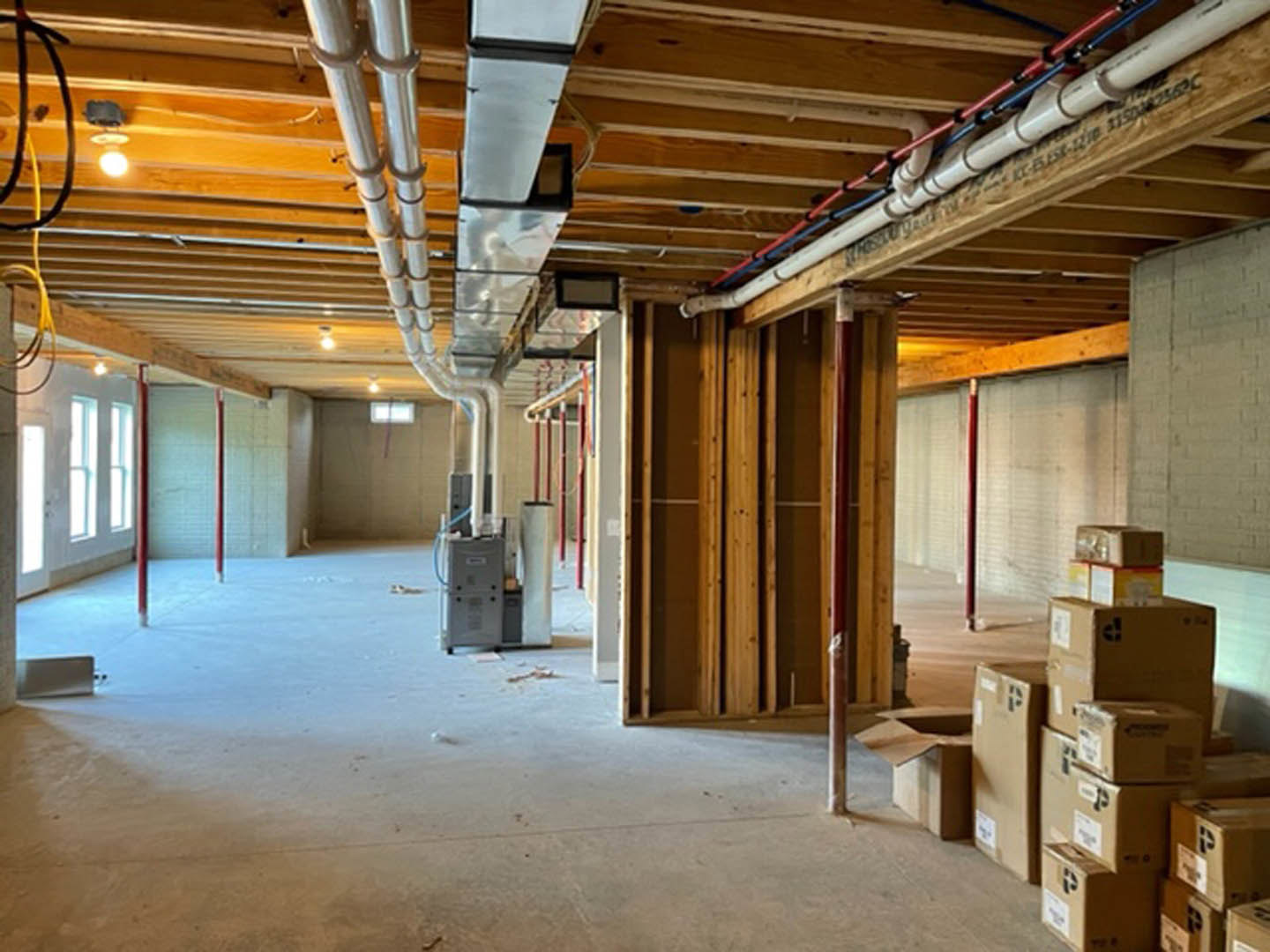 Unfinished room with exposed ceiling pipes, white walls, pile of cardboard boxes on the floor, and visible beams