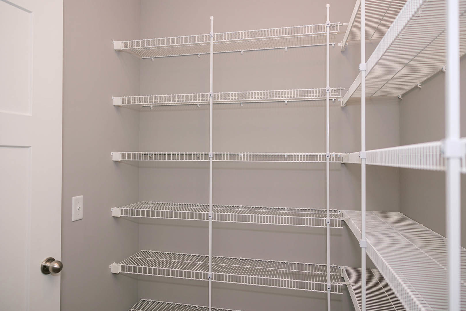 White wire shelving with metal rods mounted on a wall, adjacent to a white door with a close-up of a silver door knob and a white light switch, in a bright indoor room.