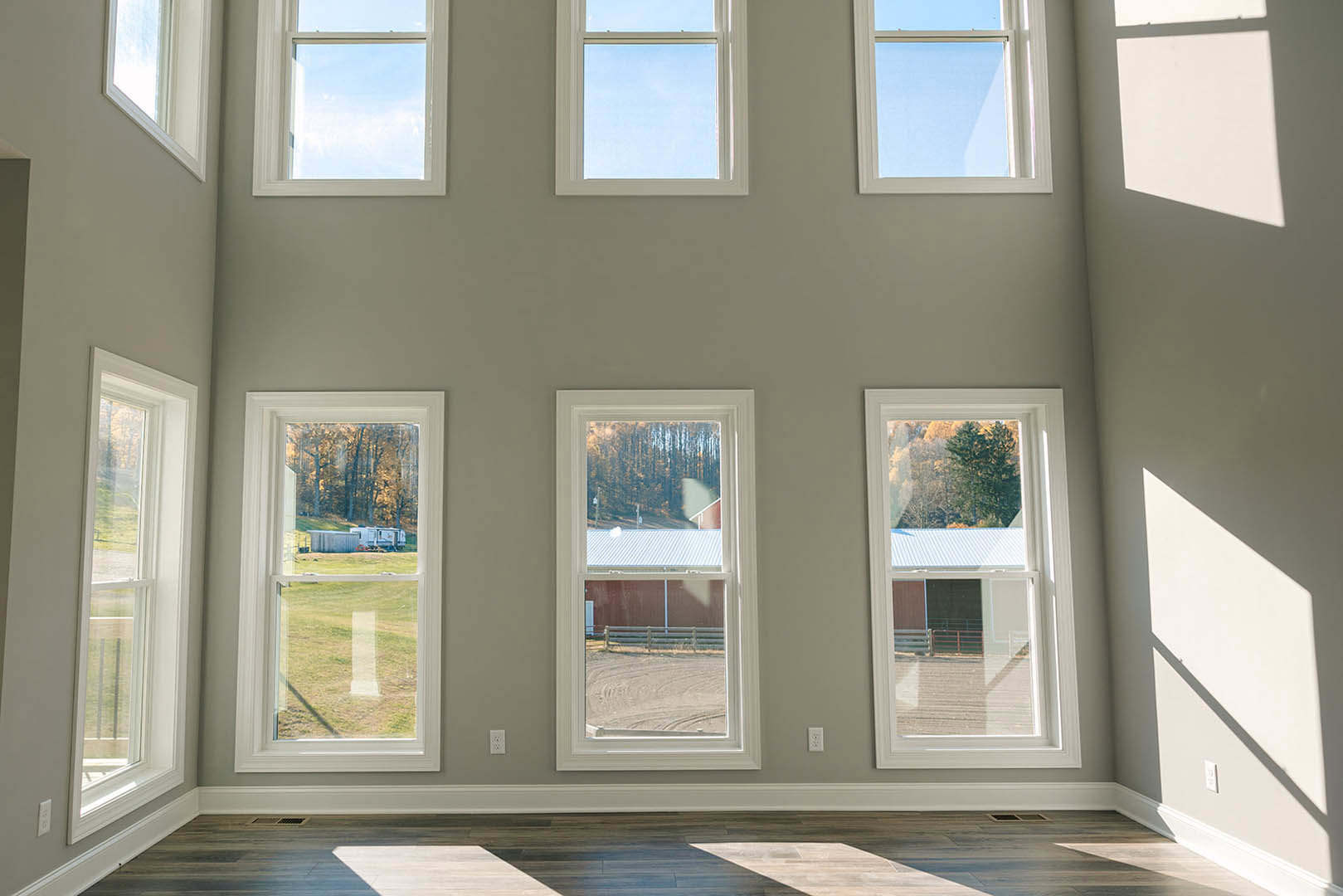 Spacious room featuring multiple large windows, wooden flooring, and views of a barn, trailer, and farm under a clear sky.