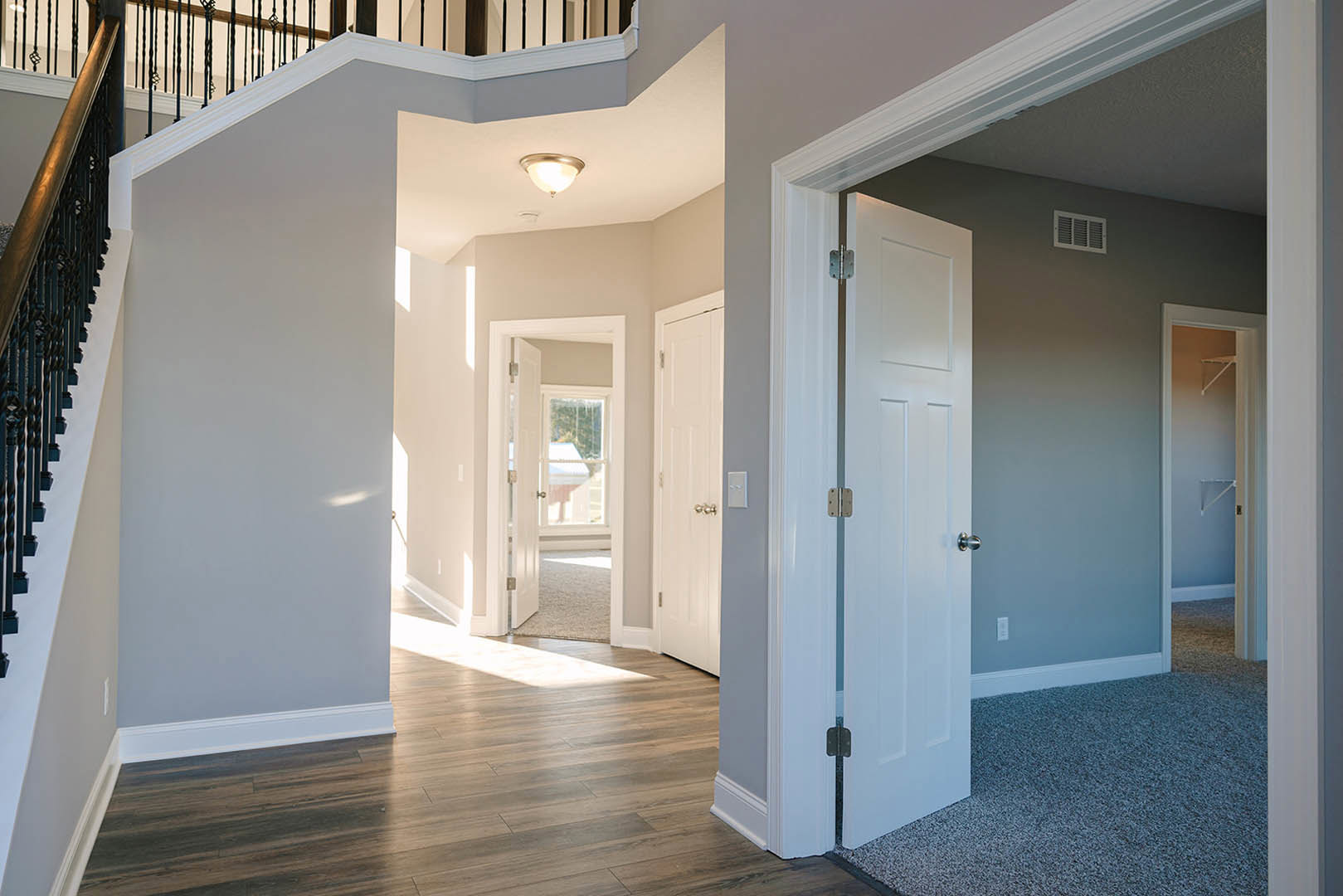 Wide hallway with hardwood flooring, white walls, and a staircase featuring a metal handrail; multiple white doors, one with a window, and a ceiling light fixture illuminated.
