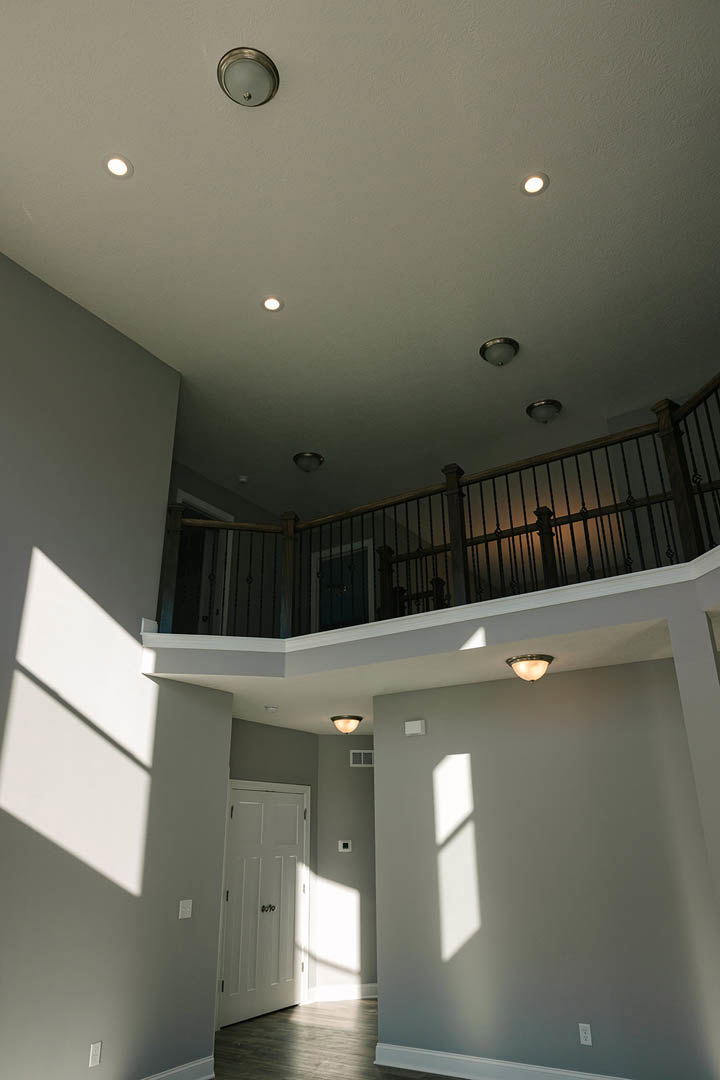 Dark wood flooring and white walls in a bright room, featuring a white door with black knobs, a balcony with metal railing, and sunlight casting window shadows across the floor.