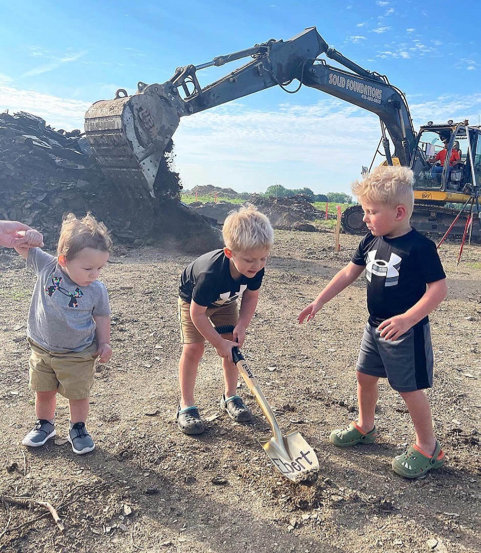 Children playing and digging in a dirt field near a large excavator, surrounded by mountains and cloudy sky, wearing casual clothing and footwear