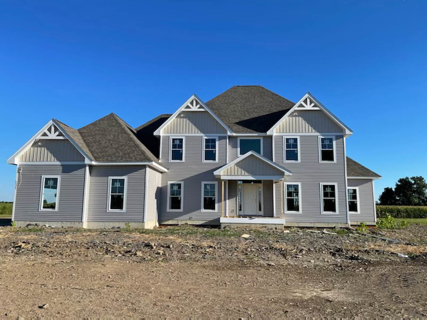 Two-story farmhouse under construction with white siding, white-framed windows, front porch, dirt yard, scattered grass and rocks, blue sky overhead