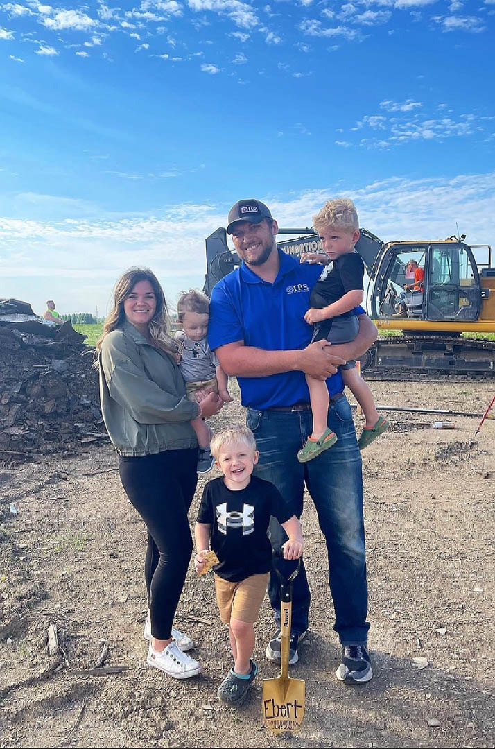 Group of adults and children standing on a dirt lot in front of a partially constructed custom home with exposed framing, wearing casual clothing under a cloudy sky