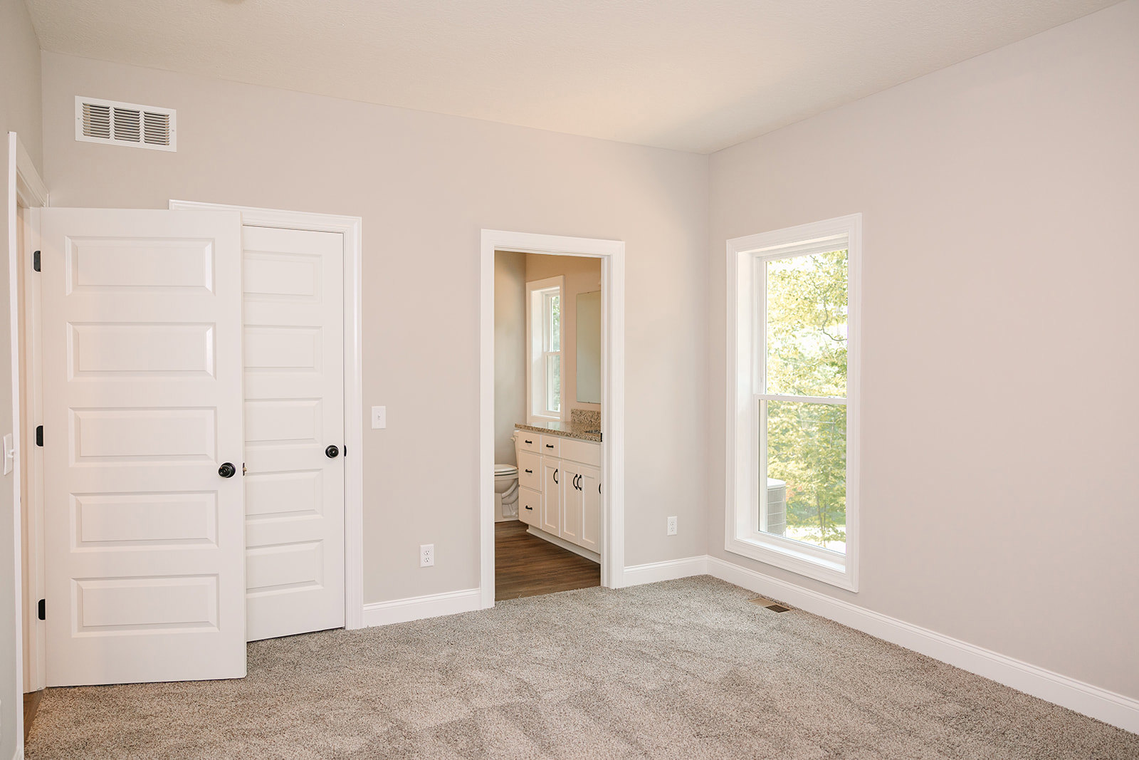Bedroom with light carpet flooring, two white paneled doors with black knobs, ceiling vent, window overlooking trees, and adjacent bathroom entrance