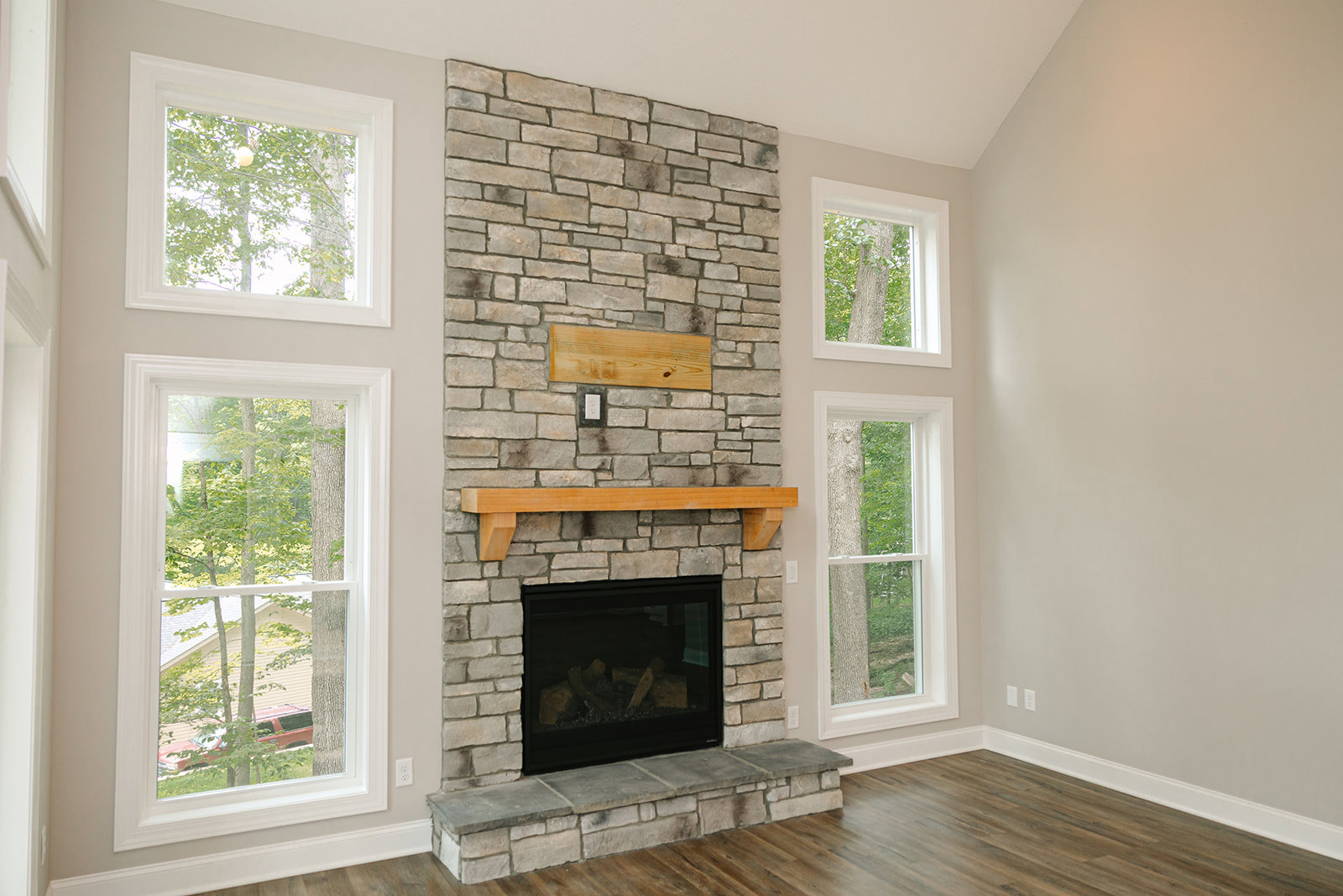 Stone fireplace with stacked logs, wood mantel shelf, hardwood flooring, large window framing leafy trees, textured stone wall accents
