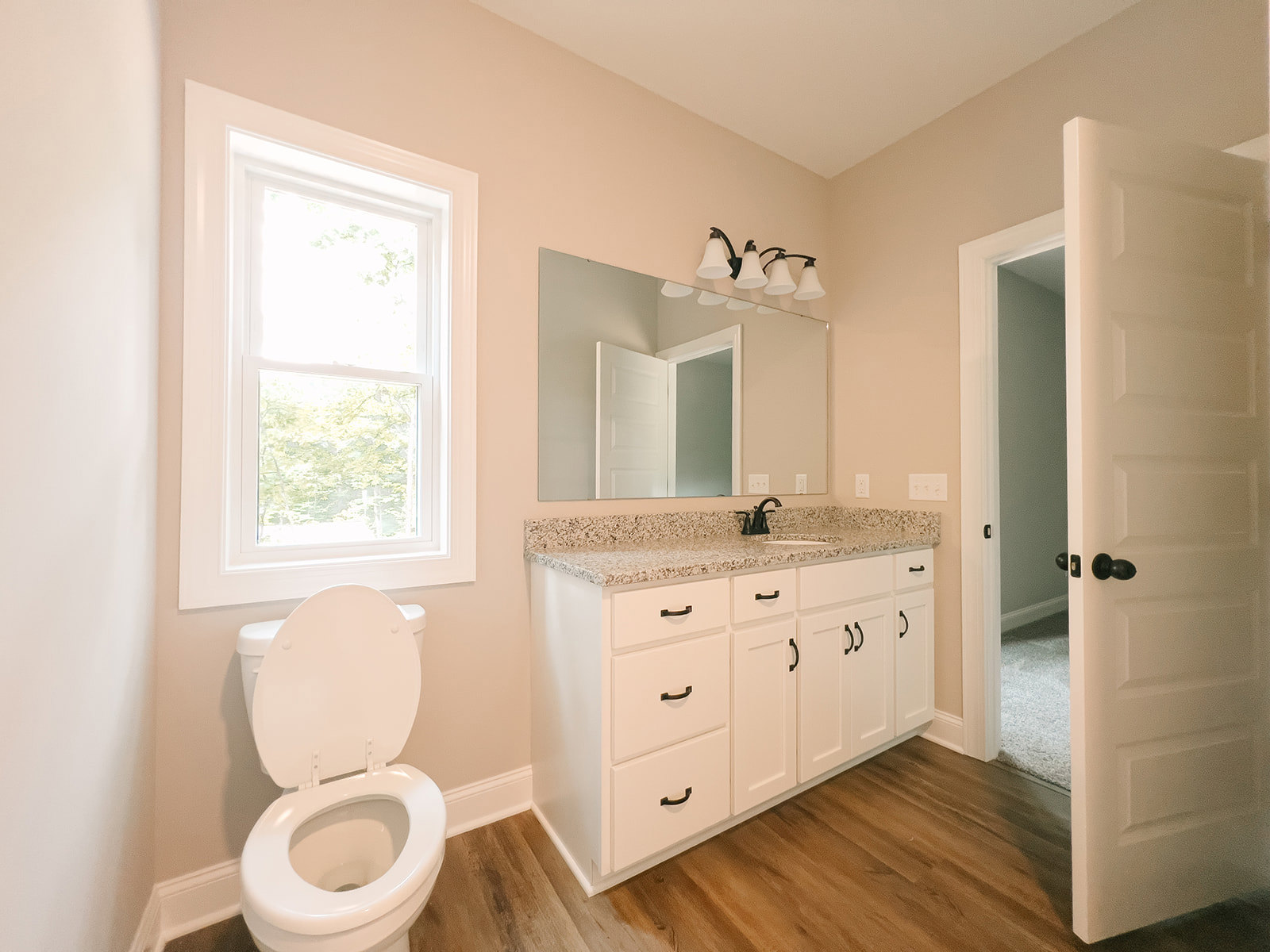 Bathroom with white toilet, white sink, white-framed window, white door with black knobs, wall-mounted light fixture, neutral tile flooring, and wall-mounted mirror above the sink.