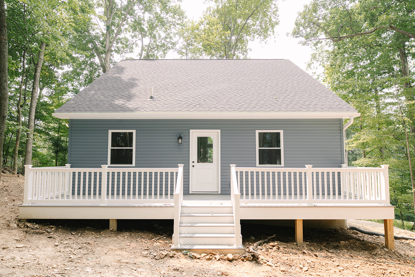 Front porch with white stairs and railing, white door with window, white-framed window, gray siding, gabled roof, small tree and plants along entry walkway