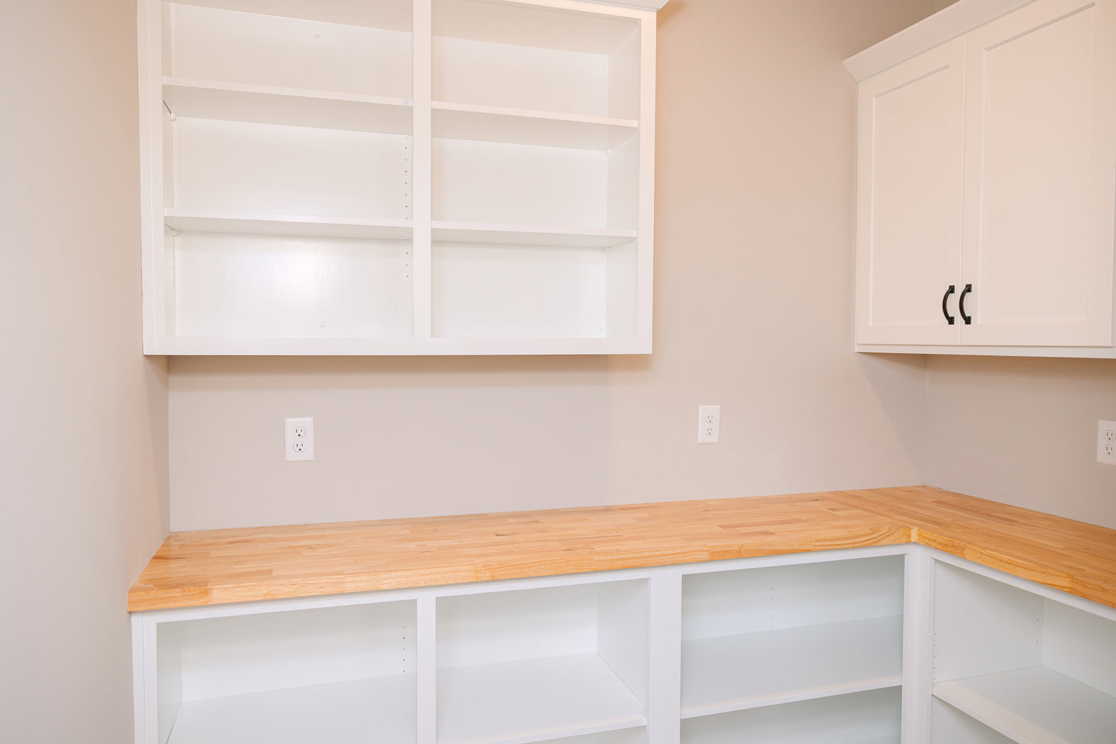 White kitchen shelves with wooden countertop, white cabinets featuring black handles, electrical outlet visible on backsplash, light plywood accents.
