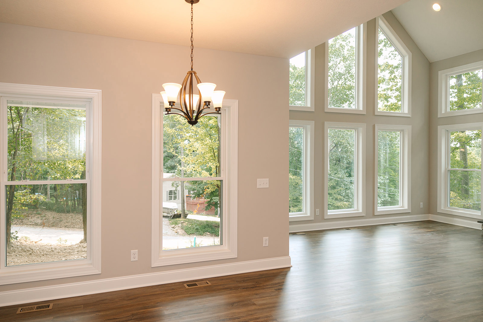 Spacious room with large windows overlooking trees, wood flooring, white ceiling with decorative chandelier, and crown molding accents