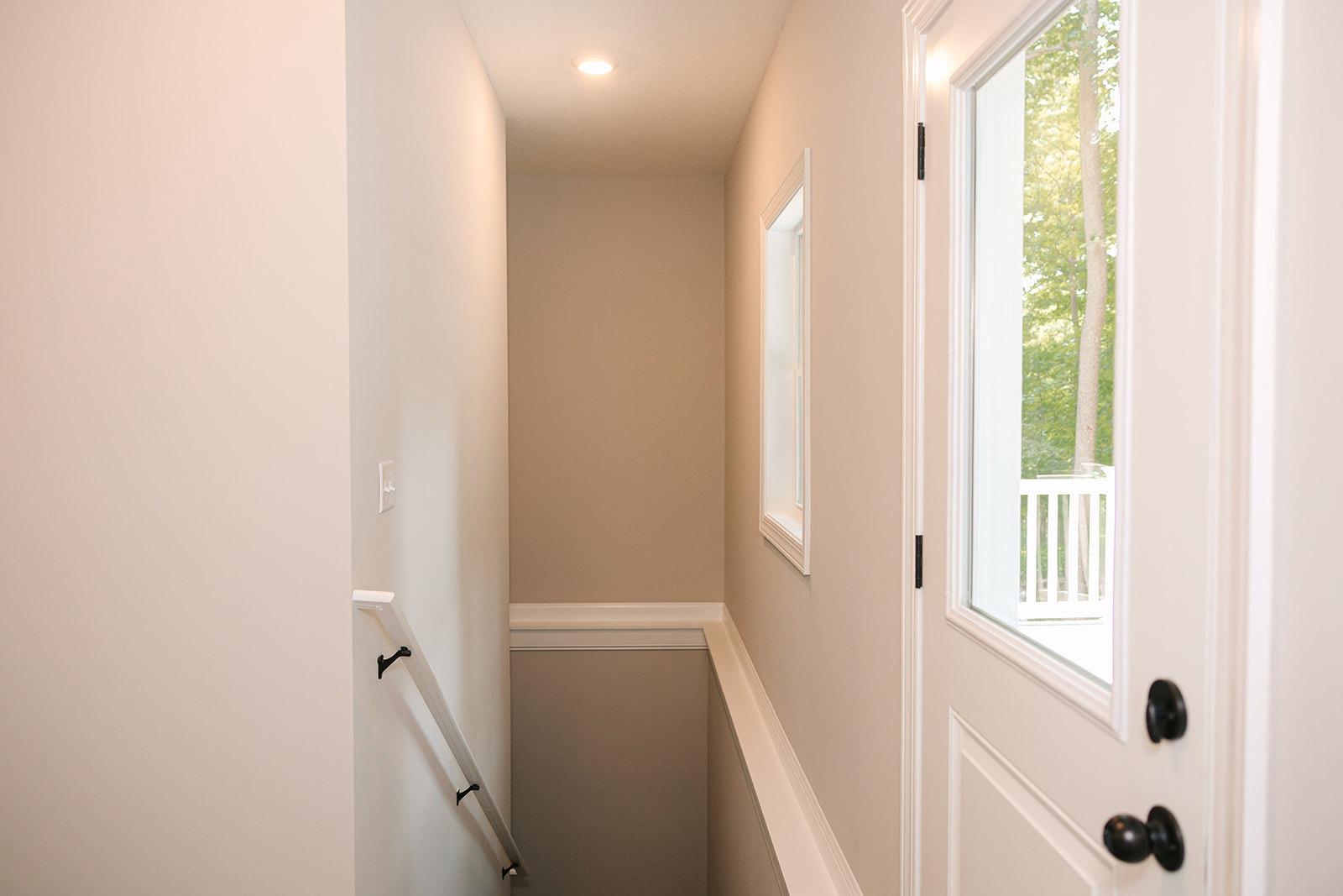 Hallway with white walls, black metal staircase railing, white door with central light fixture, window showing trees outside, grey accent wall