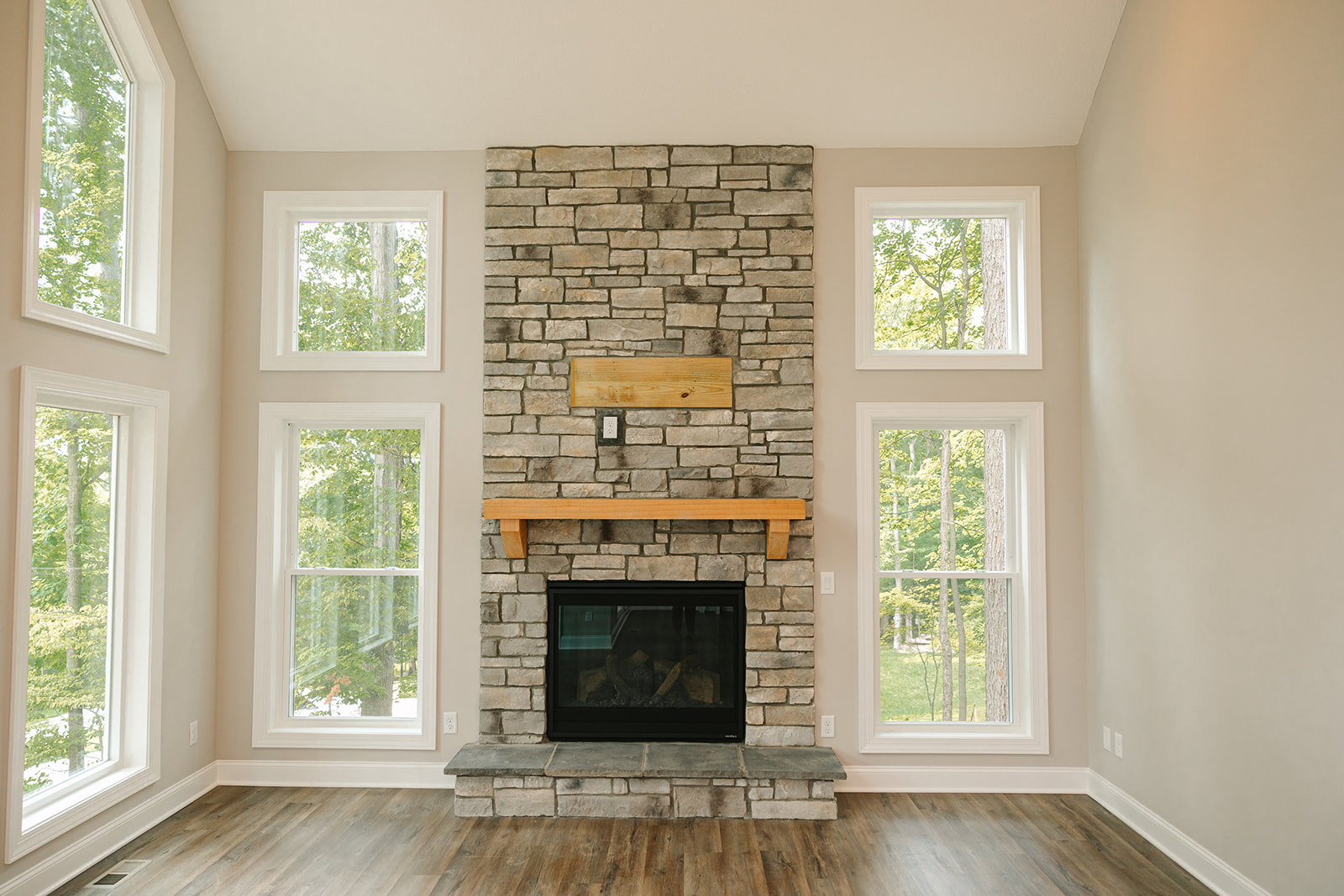 Stone fireplace with wood mantel, stacked firewood, large windows framing tree views, hardwood floors, neutral walls
