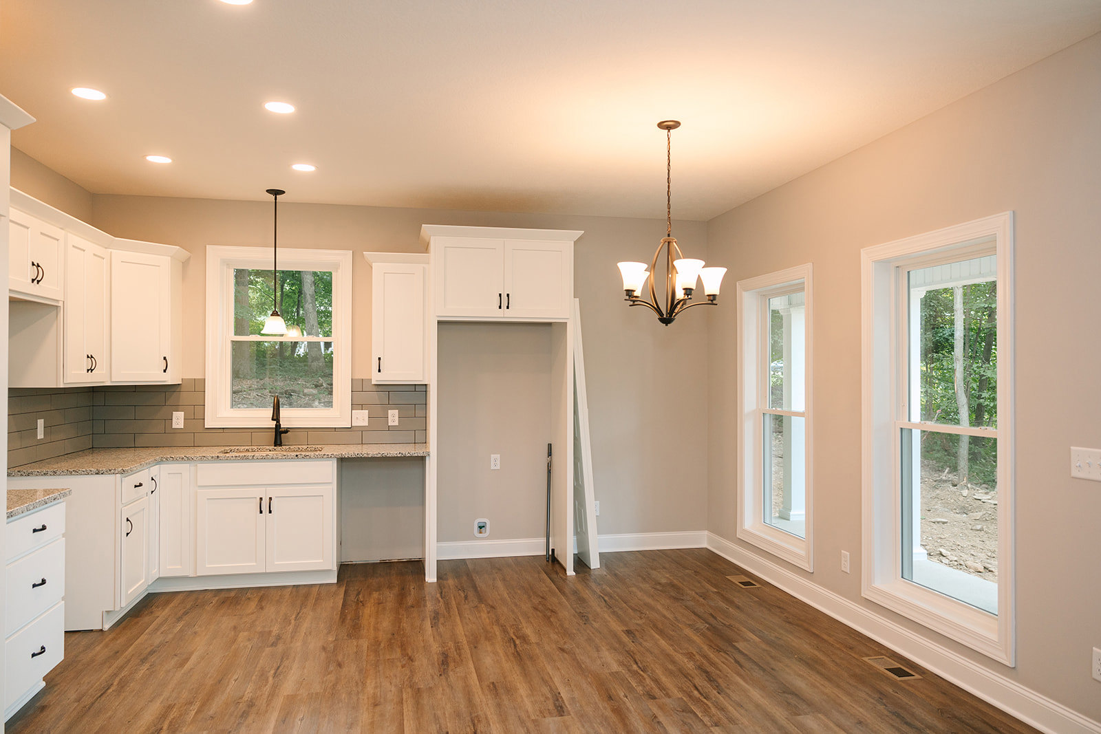 Kitchen with warm wood flooring, white cabinetry with black handles, modern chandelier, large window overlooking trees, and ceiling light fixture.