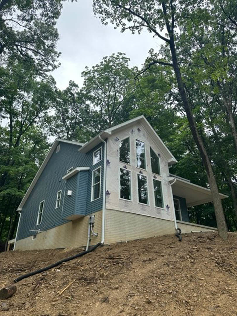 Two-story home with light siding and large windows set on a grassy hill, surrounded by mature trees under a clear sky