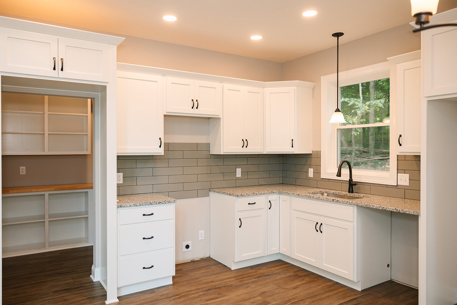 Kitchen with white shaker cabinets, granite countertops, black hardware on drawers, sunlight streaming through window, white shelving with wooden top