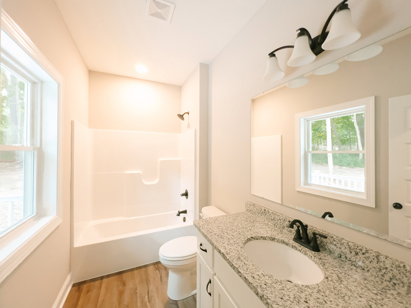Bathroom with white sink and black faucet, white toilet, bathtub, large window framed in white showing trees outside, light tile walls and floor, rectangular mirror above sink.