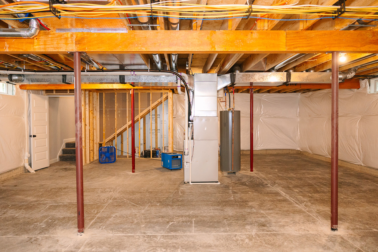 Unfinished basement with exposed steel beams, white insulated walls, visible wiring, metal utility box, blue ventilation fan, and pipes along the ceiling and floor.