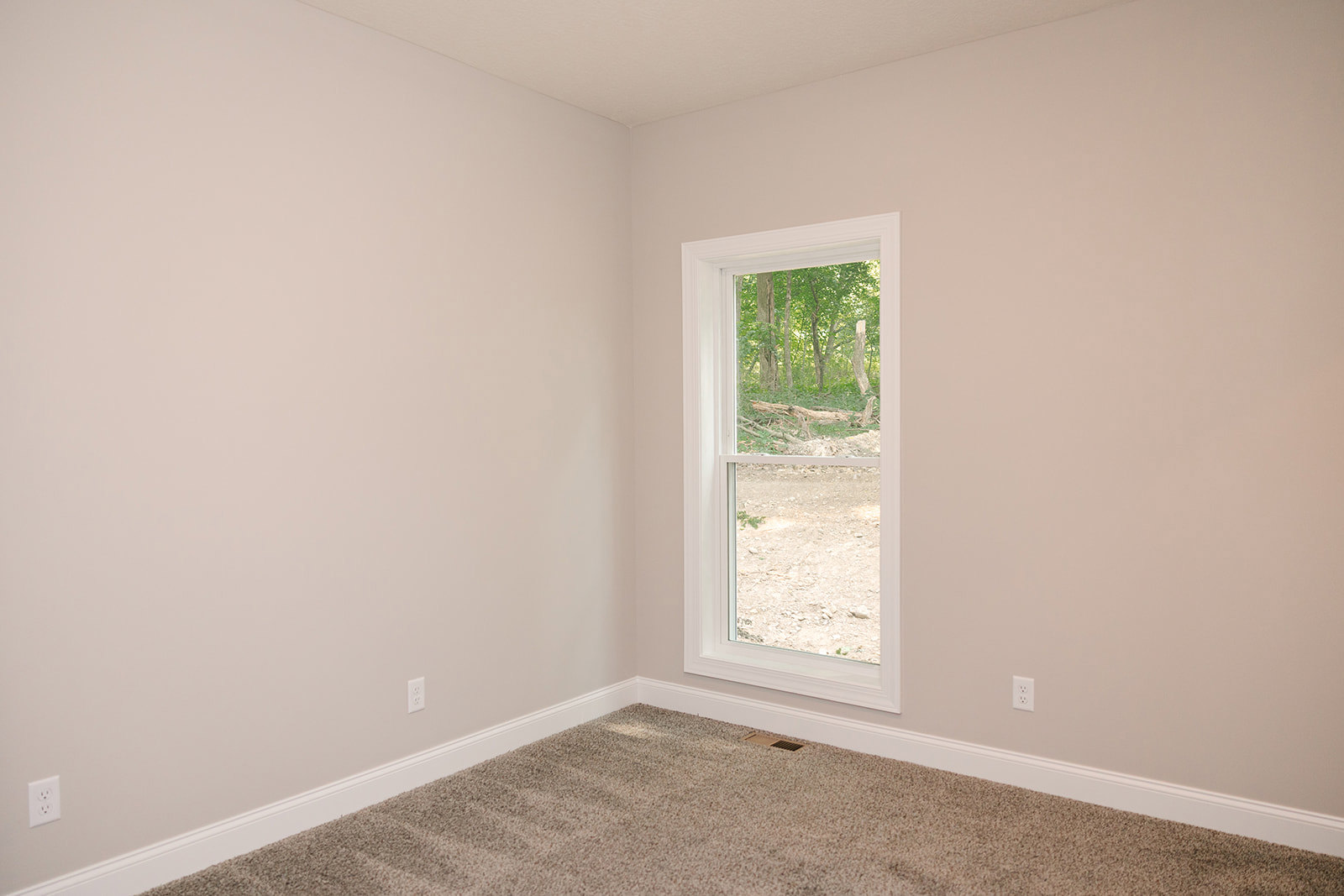 Neutral-toned carpeted bedroom with white walls, large window overlooking dense forest, ceiling vent, electrical outlet, and plaster ceiling.