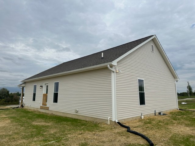 Grey-roofed house with light siding, grassy lawn, black hose and white pipe near foundation, cloudy sky overhead