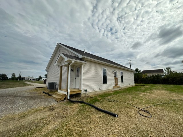 Two-story house with white siding, large windows, and a garden hose extending across the front lawn; surrounded by grass, trees, and a partly cloudy sky.