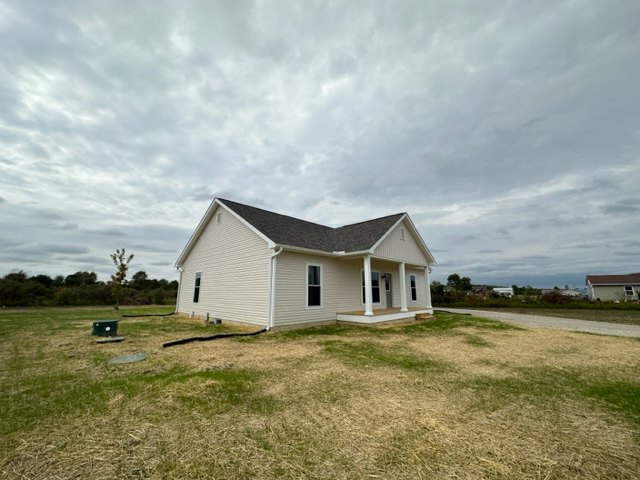 White farmhouse with covered porch, gray roof, and hose attached to side, surrounded by grassy lawn under cloudy sky