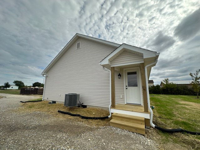 White mobile home with grey siding, white door featuring a window, wooden porch, garden hose coiled near entrance, grassy yard, cloudy sky overhead.